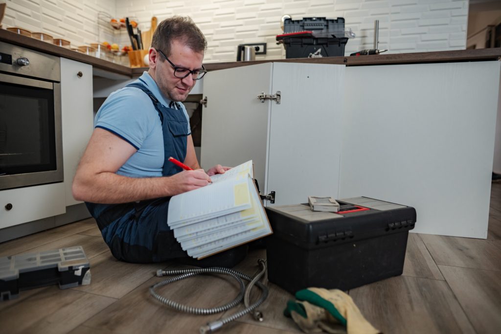 An appliance technician kneeling on a kitchen floor, writing up a job on a clipboard beside an open cabinet and a tool box