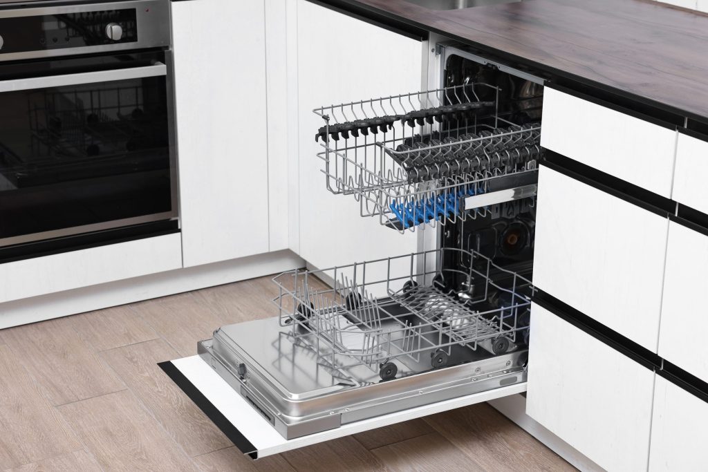 An integrated dishwasher standing open in a white handleless kitchen, empty racks pulled out, a wall oven column beside it