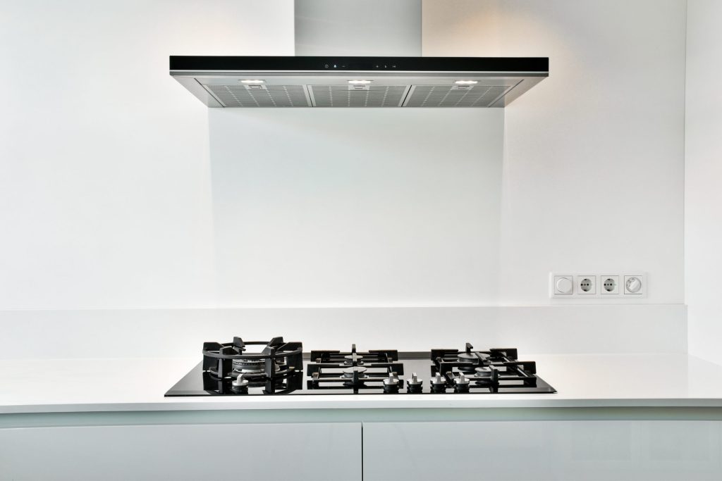 A wall-mounted chimney range hood with a black glass fascia above a gas cooktop in a white kitchen