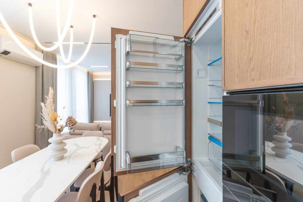 A built-in refrigerator standing open in a warm modern kitchen, its glass shelves empty