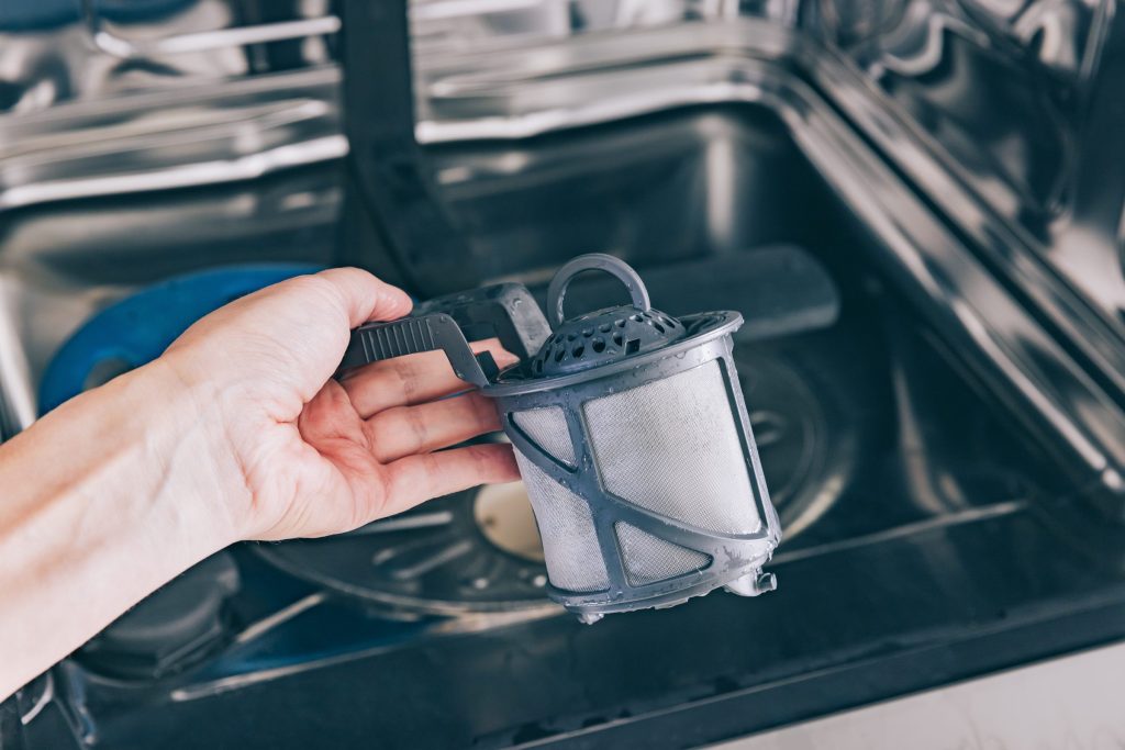 A hand lifting the cylindrical filter out of a dishwasher, the empty filter well below it