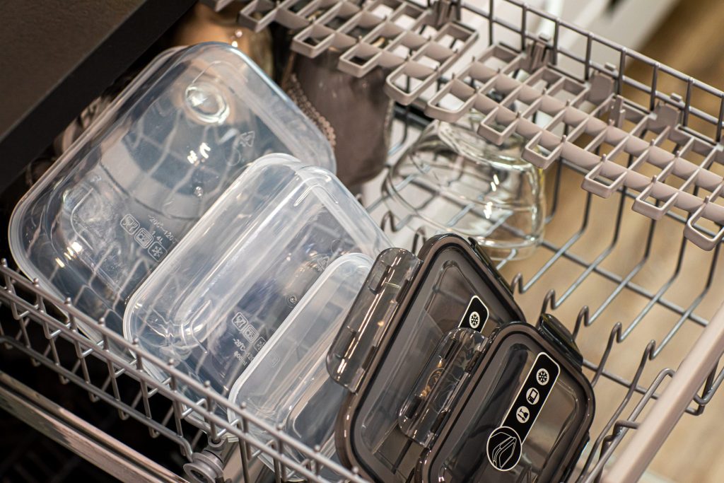 Clear plastic food containers standing in the rack of a dishwasher, water still beaded on them