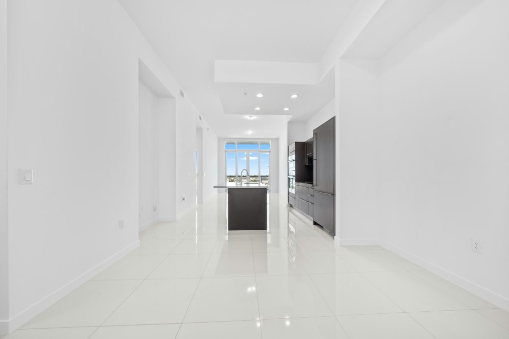 An empty white apartment with a kitchen island at the far end and the sky beyond