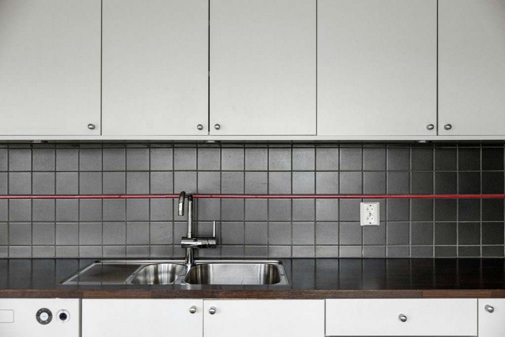 A double sink and tap against dark square tile between white cabinets