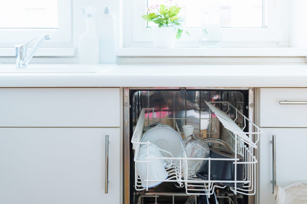 A dishwasher standing open under a bright window, clean dishware in the upper rack