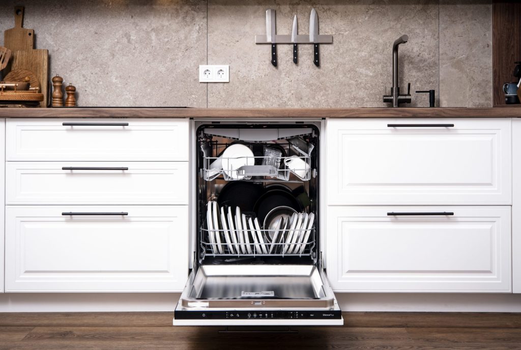 A dishwasher loaded with clean plates and glasses, set between white cabinets under a stone splashback