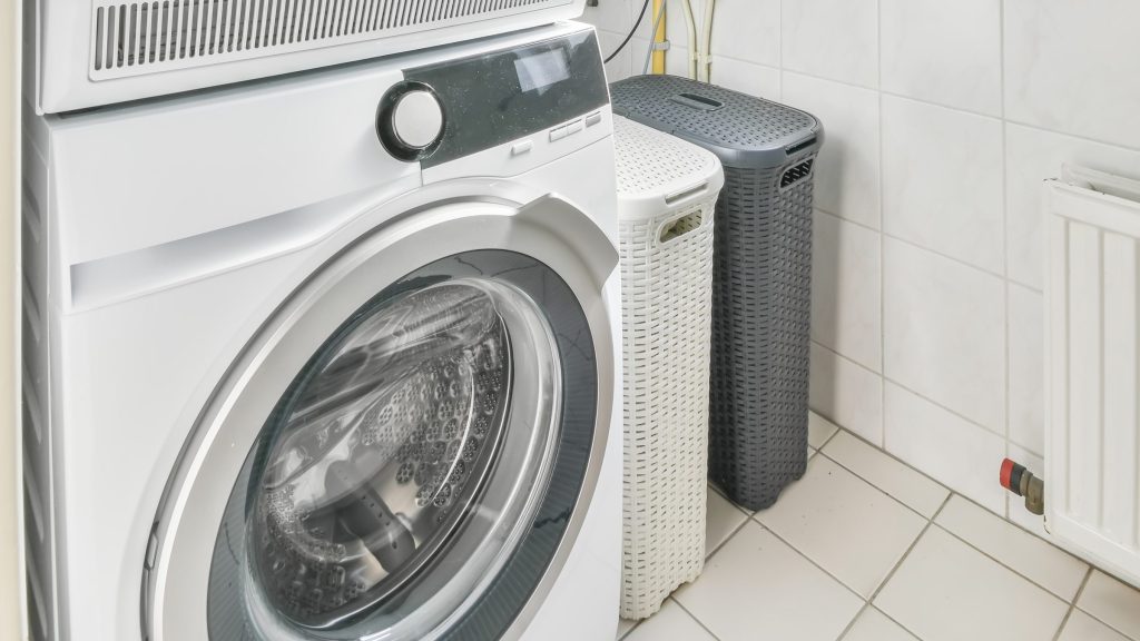A compact front-loading washer with a dryer stacked above it, in a tiled corner beside two laundry baskets