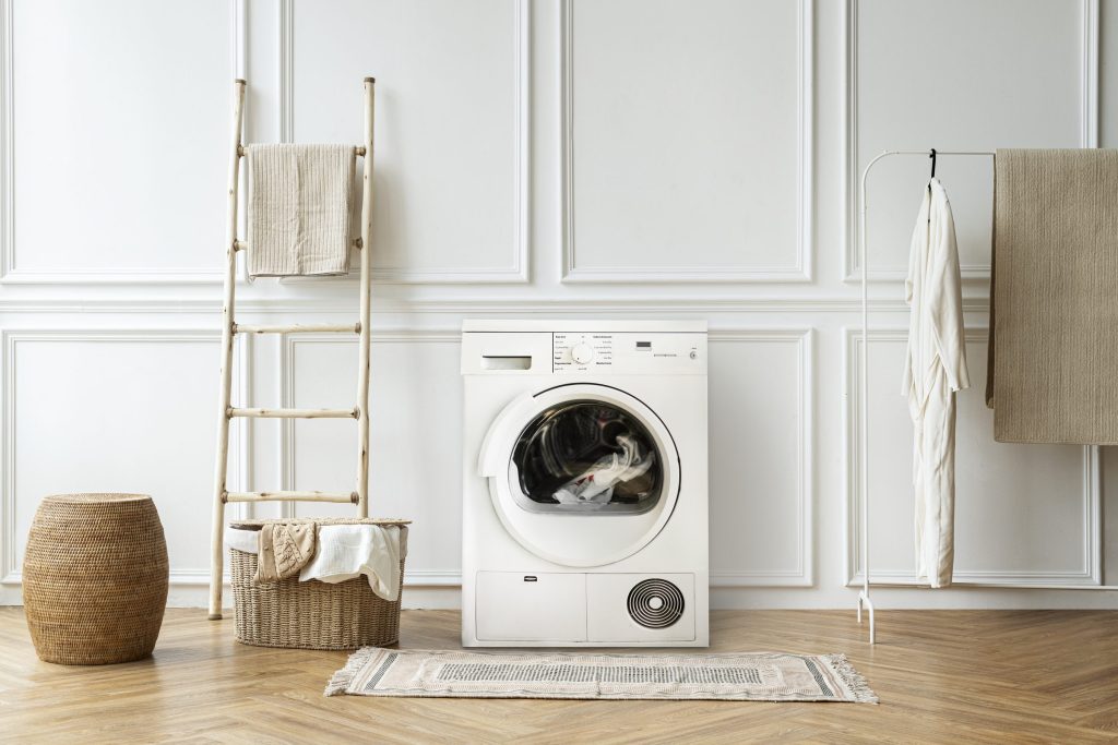 A white condenser dryer with laundry in the drum, standing against panelled walls beside a towel ladder