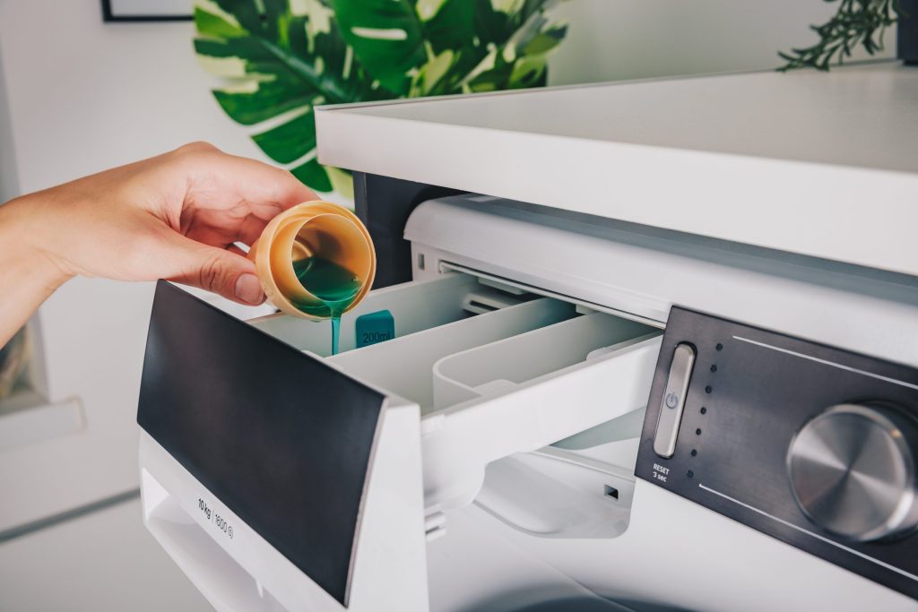 A hand pouring liquid detergent into the open dispenser drawer of a washing machine