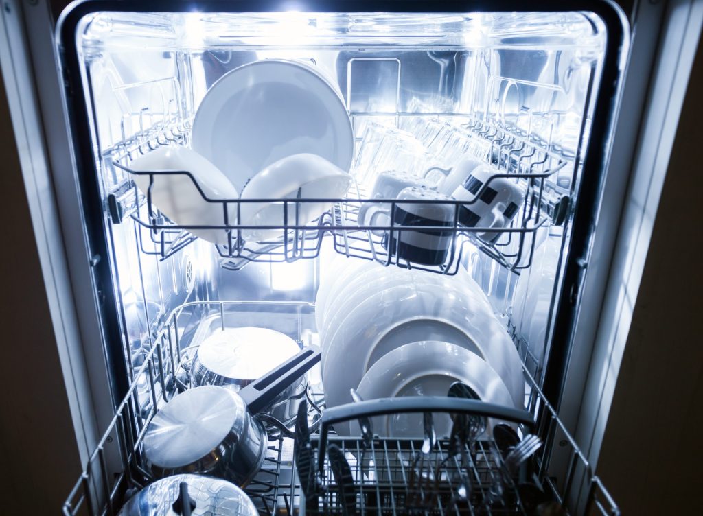 The lit interior of an open dishwasher with clean plates, cups and cutlery on both racks