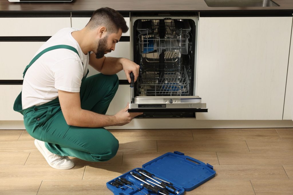 A technician kneeling at an open built-in dishwasher with an open tool case on the kitchen floor