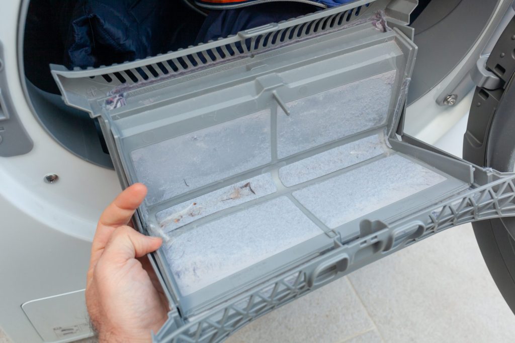 A hand holding open the lint filter of a white dryer, the mesh grey with collected lint