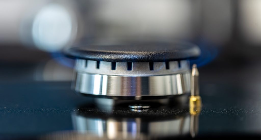 A gas burner cap photographed close up with the spark igniter electrode beside it and a blue flame behind