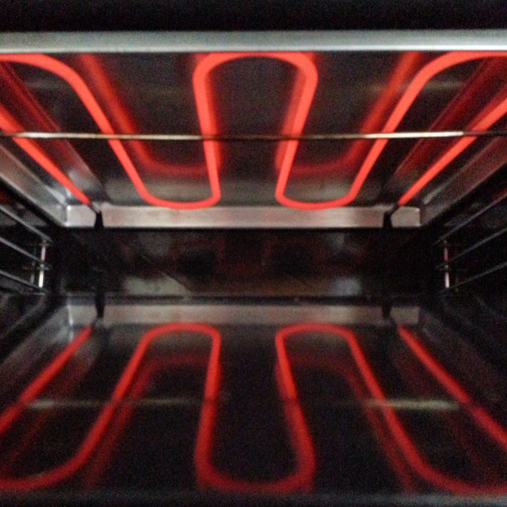 The grill element glowing red across the roof of an oven cavity, reflected in the steel walls