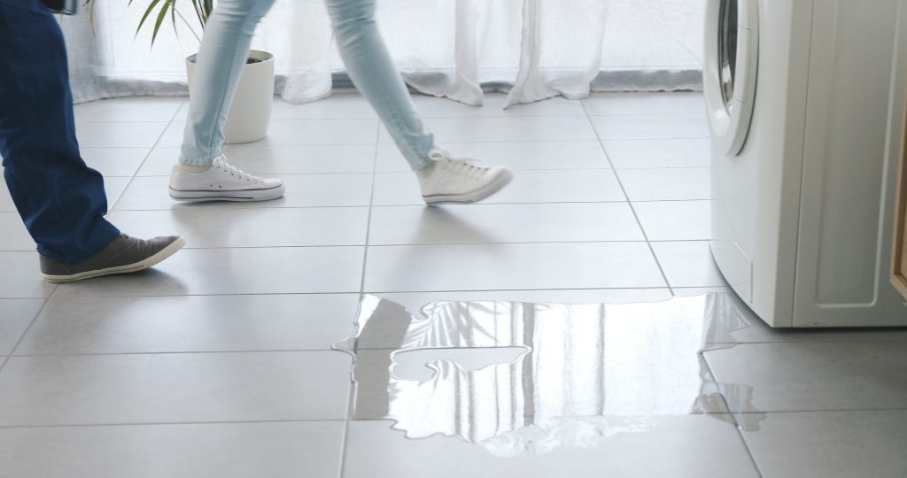 A puddle of water spreading across a tiled floor in front of a washing machine