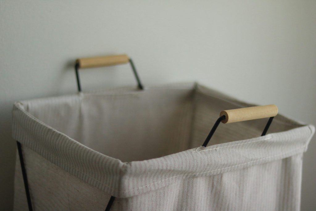 An empty canvas laundry hamper with wooden handles against a pale wall