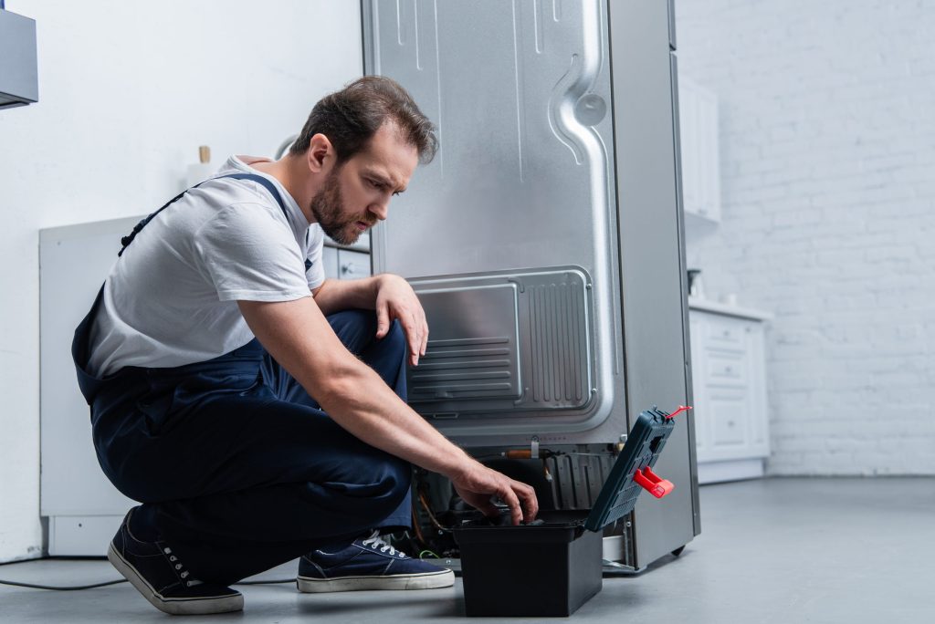 A technician crouching behind a stainless refrigerator pulled out from the wall, reaching into the open toolbox at its foot